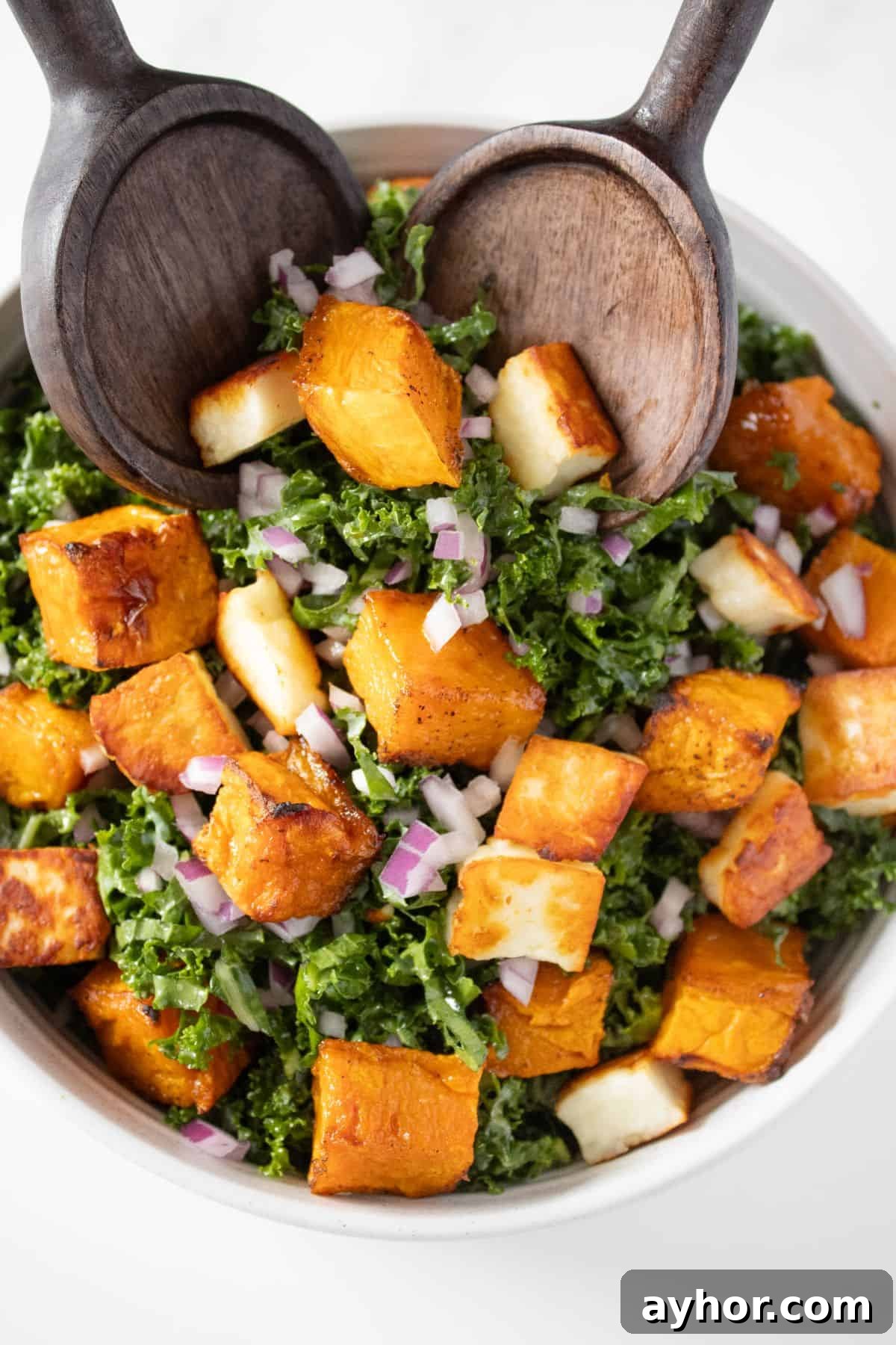 Round white bowl of Kale and Pumpkin Salad, with some wooden serving utensils scooping up some salad.