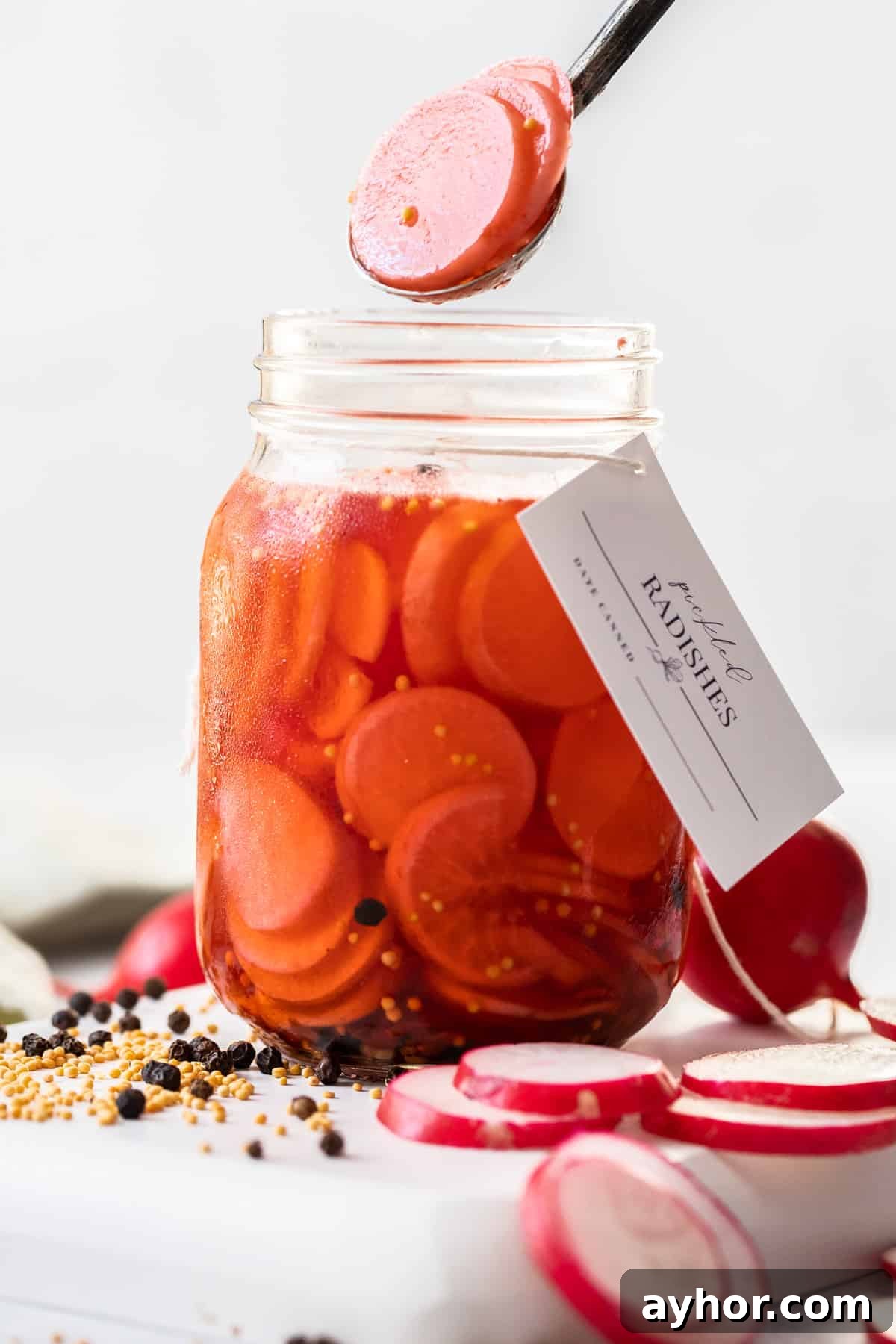 Glass jar of Pickled Radishes, with a label around the neck of the jar and a spoon lifting out some radish slices.