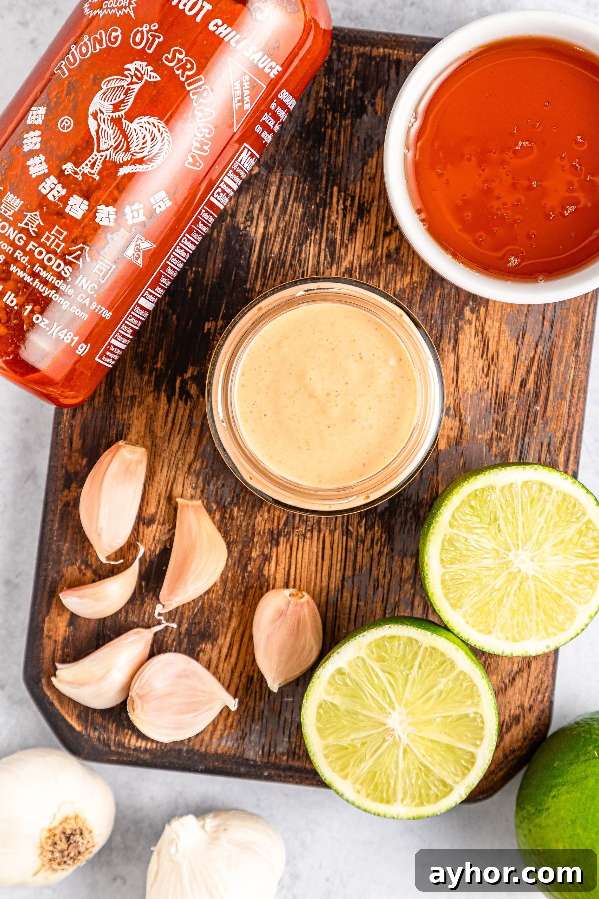 Recipe for Sriracha Aioli (Gluten-Free, Dairy-Free) Garlic cloves, lime wedges, Sriracha and sriracha aioli on wooden board.